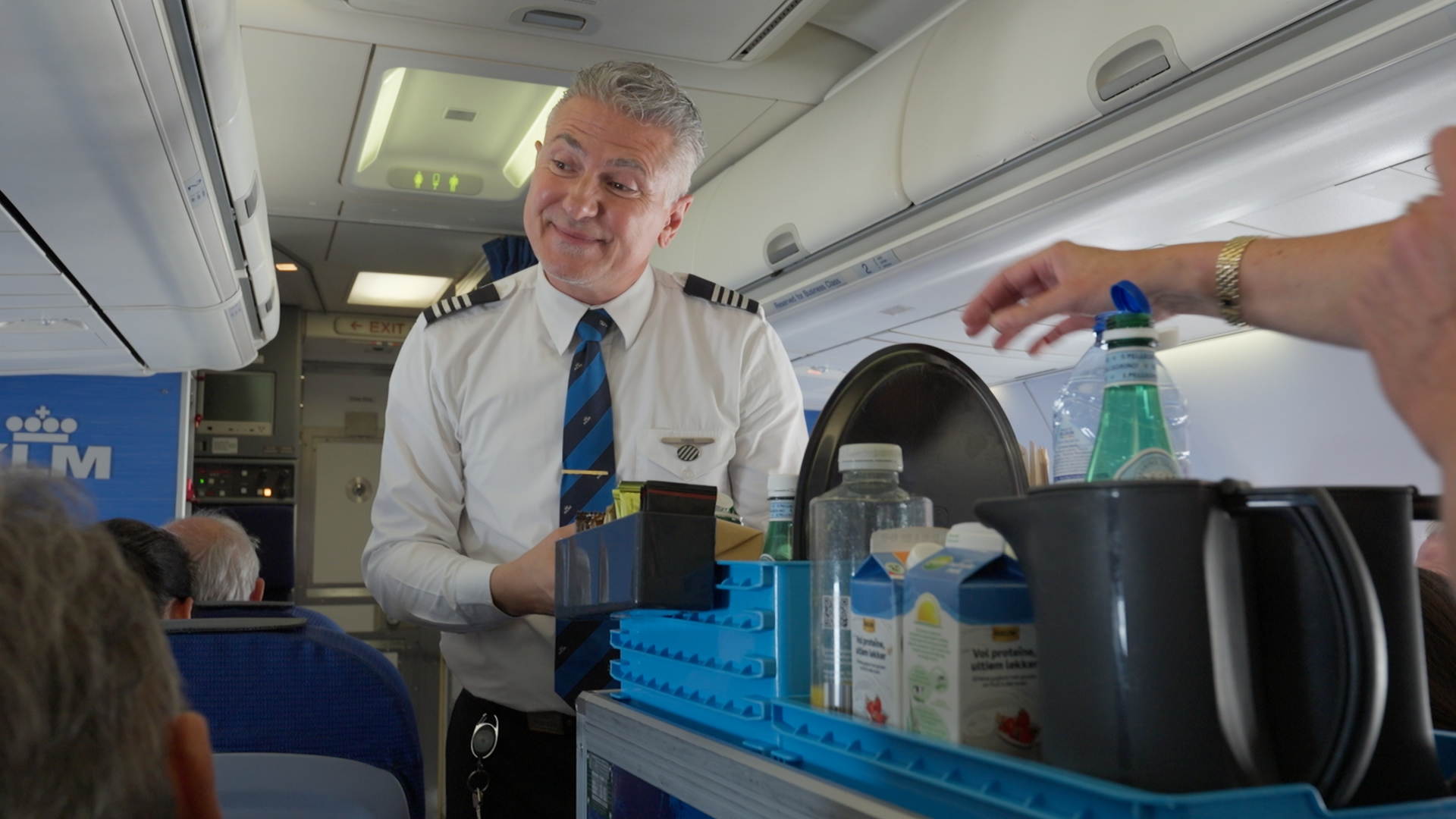Een KLM-cabinemedewerker in wit overhemd met stropdas bij een trolley met dranken in het gangpad van een vliegtuig