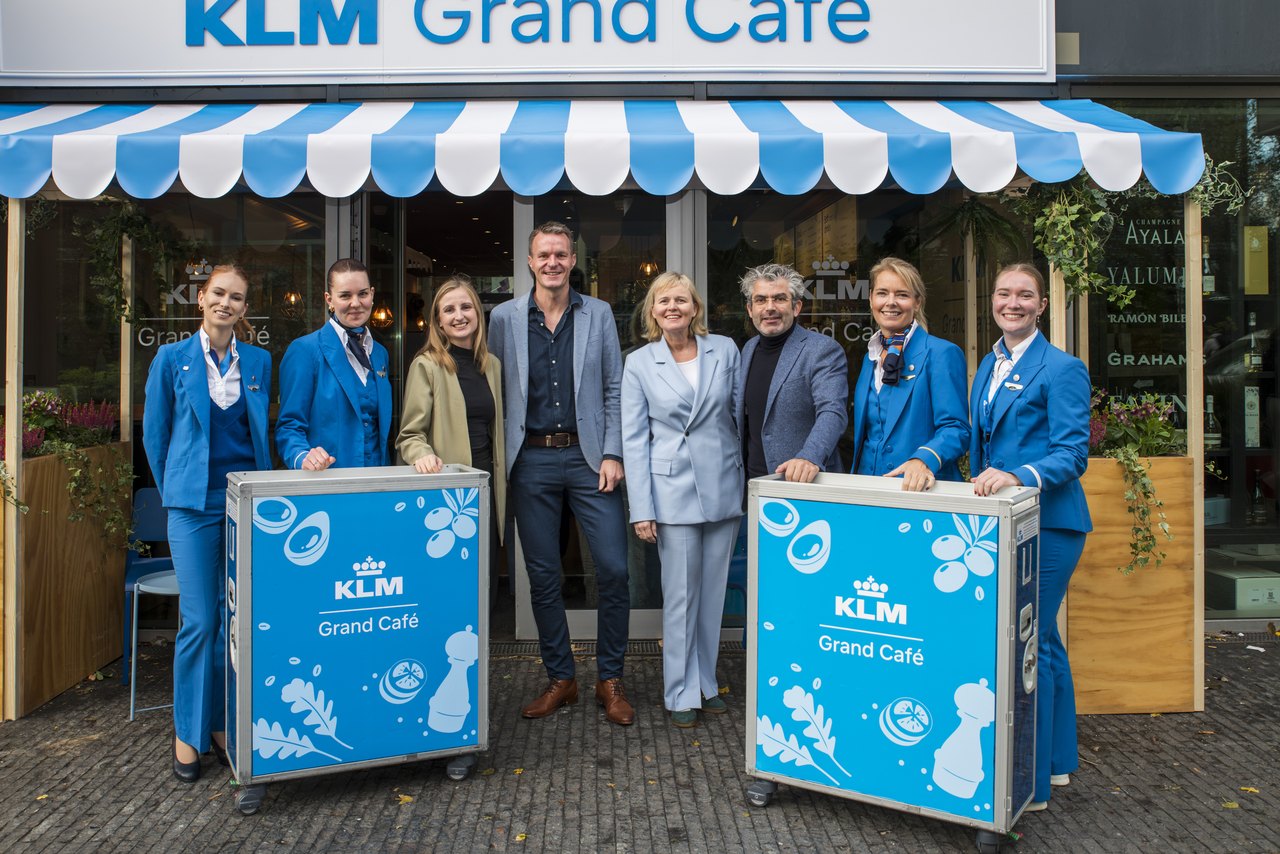 Cabinepersoneel en medewerkers van KLM bij twee blauwe trolleys met het opschrift Grand Café voor het tijdelijke café in Amsterdam Zuid