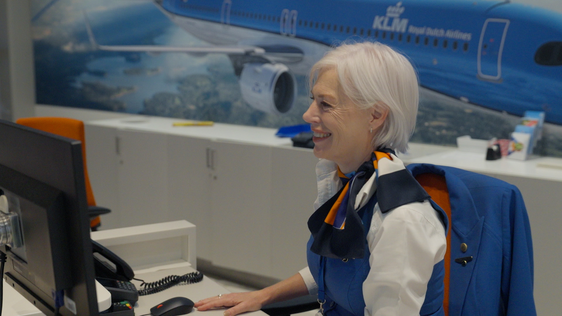 Een KLM-medewerker in uniform met sjaal achter een balie met een computerscherm, met een grote foto van een KLM-vliegtuig aan de wand