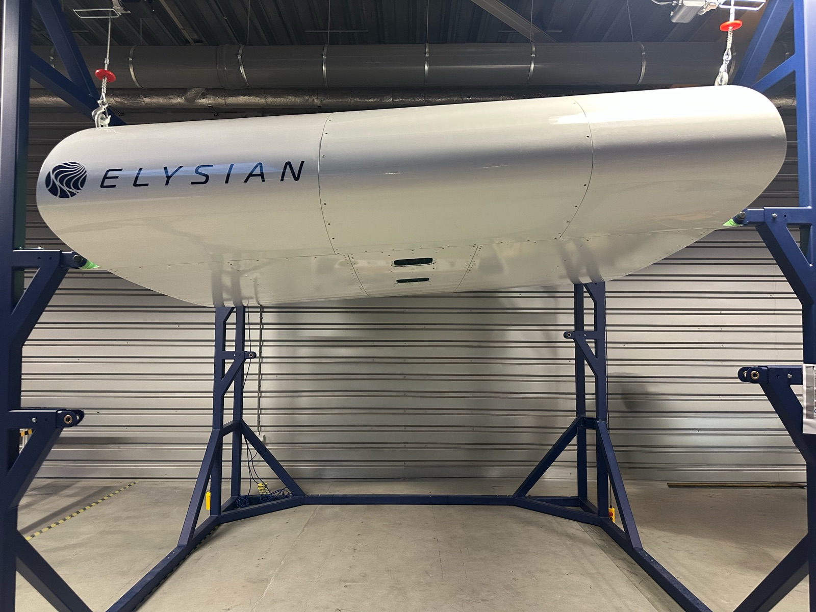Een wit model op ware grootte van een deel van de vleugel van de E9X met het logo van Elysian, opgehangen in een blauw stalen frame in een werkplaats