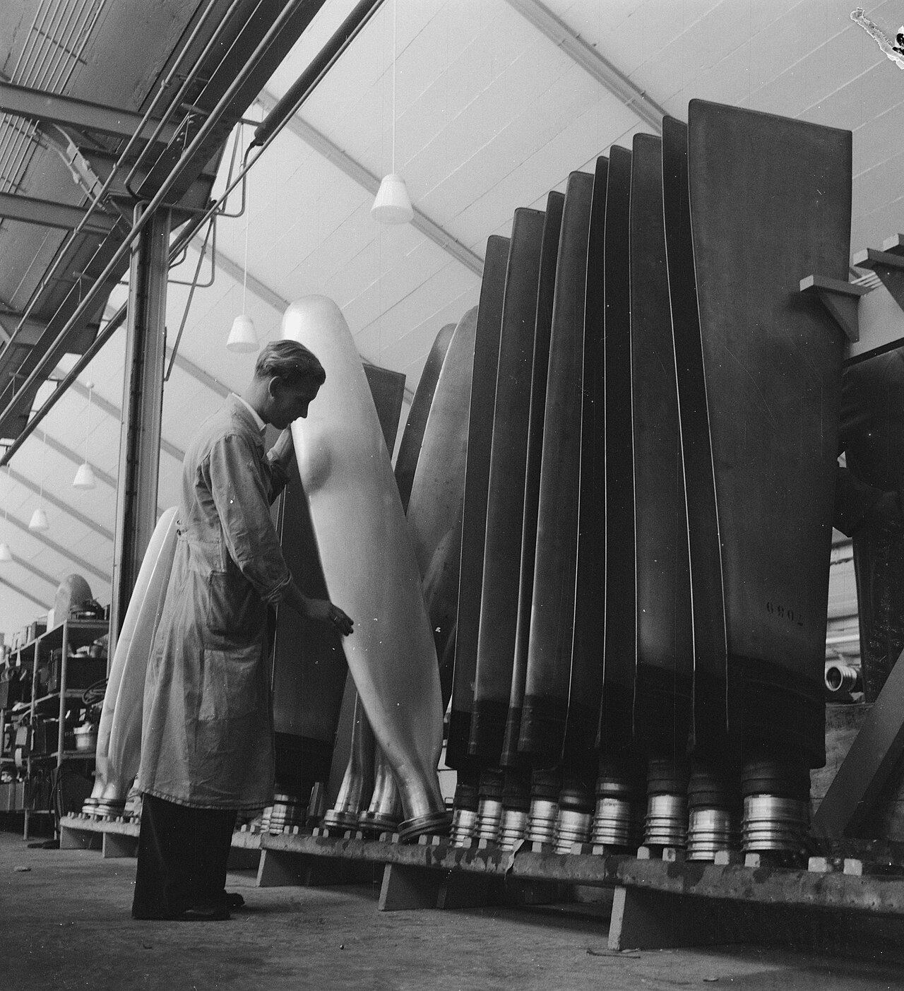 Zwart-witfoto uit 1951: een monteur in een lange stofjas staat tussen een rij grote propellerbladen in een werkplaats van de technische dienst van KLM op Schiphol.