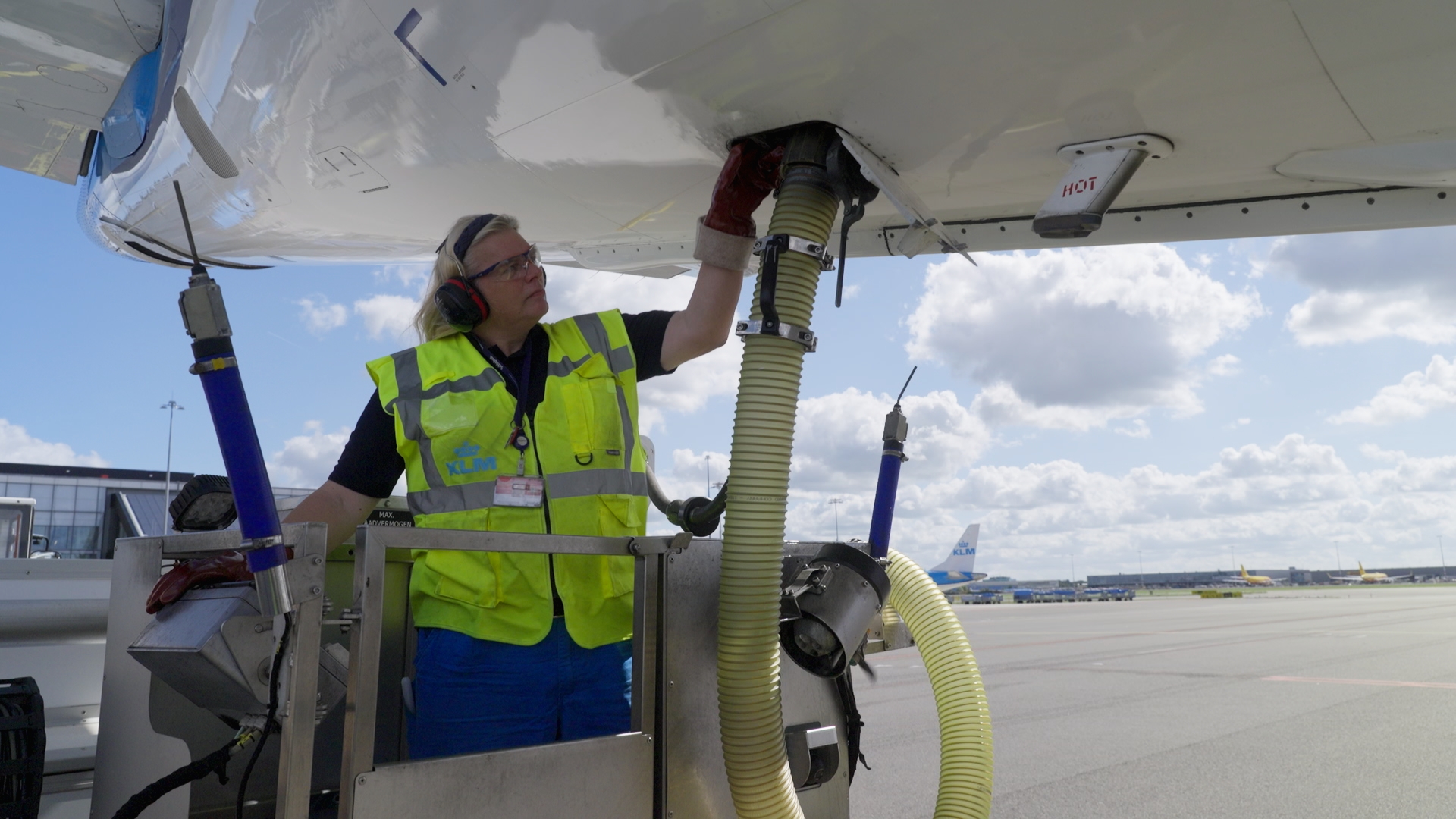 Een KLM-medewerker met gehoorbescherming en een geel hesje werkt staand op een servicewagen onder de romp van een vliegtuig, met slangen naar het toestel